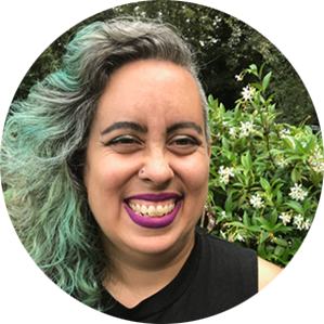 Wearing a sleeveless black t-shirt, a Sri Lankan, Roma and Irish nonbinary femme with tan skin, curly shoulder-length turquoise and grey hair, and purple lipstick, smiles brightly at the camera in front of a blooming jasmine vine.