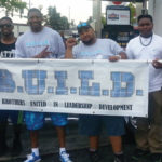 six Black men and one black child, standing outside and holding a banner that reads "B.U.I.L.D. Brothers United in Leadership Development"