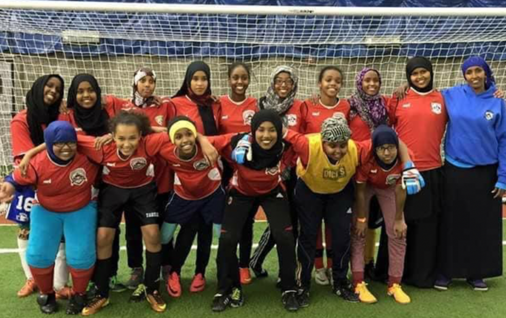 Female participants in the Somali Youth Soccer Association stand with arms around each other in front of a soccer goal.