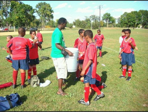 A male soccer coach carries a round cooler of water around to kids in soccer jerseys who are part of the Somali Youth Soccer Association 
