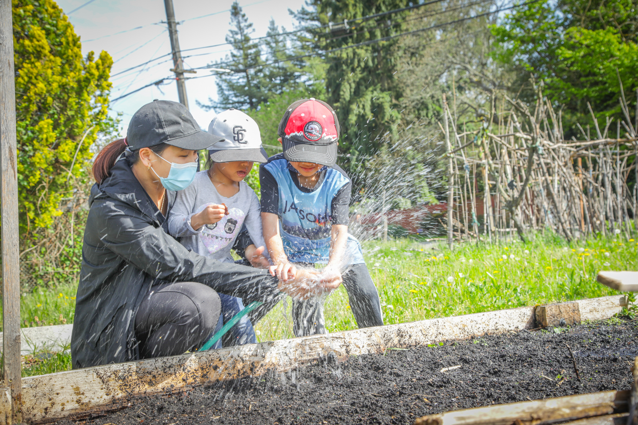 A mother wearing a baseball hat and face mask crouches next to a garden bed with her two children as they play with a spraying hose.