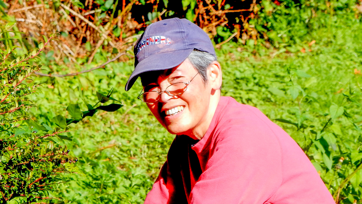 A women of Japanese descent wearing a baseball cap, sweatshirt, and gloves crouches down in a garden bed surrounded by plants.
