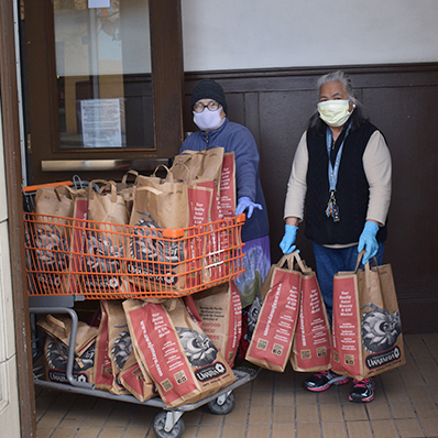two masked people rolling cart of groceries into building