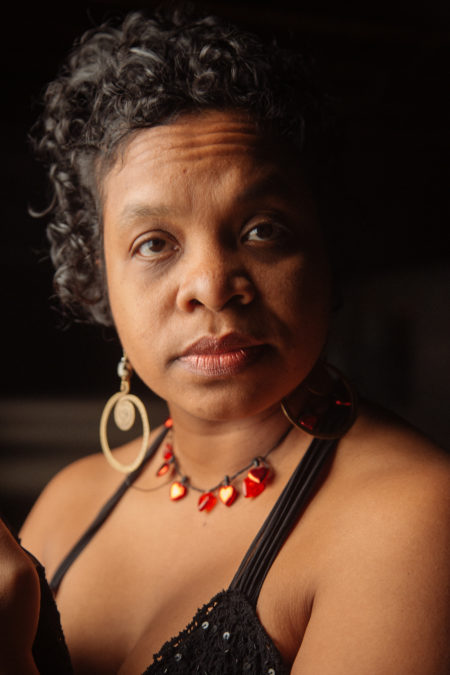 close up image of Afro Latina woman with short curly black hair, large gold earrings, and a necklace with miniature red hearts looking intently into the camera