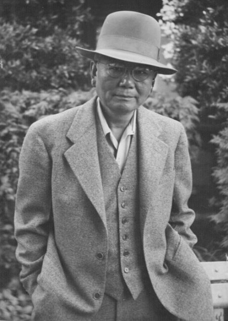 Chinese man dressed in a grey suit and hat, standing outside with his hands in his pockets. He wears glasses and is looking at the camera with a slight smile.