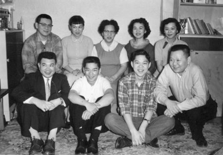 Chinese family posing for a portrait in their home. they are all sitting and smiling warmly. most are looking at the camera but a few are looking at someone or something off camera that seems to be amusing them.
