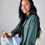 Photo of young Filipino woman in striped button-up shirt and jeans. She is sitting on a stool, holding a paintbrush, and smiling.