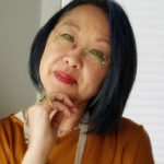 Chinese woman in glasses and orange blouse. her hand is under her chin and she is looking inquisitively at the camera.