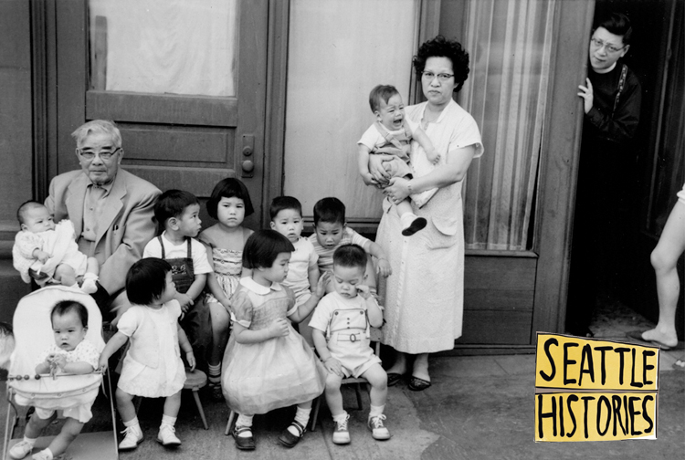 black and white photo of a group of 10 Chinese children sitting and standing on a stoop outside a building. there is an older man seated with them and an older woman standing next to them. they are each holding a baby in their arms.