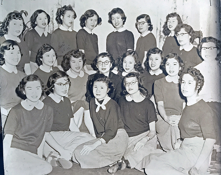 Black and white photo of a group of Chinese American girls dressed in matching sweaters with white collars and skirts. Shiny curls and lipstick, sweet smiles on all the girls all lined up in three rows.