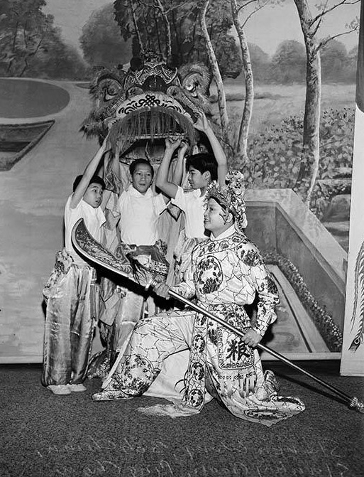 Black and white photo of three children holding up a Chinese lion mask, fronted by a teen or adult holding a scepter and wearing intricate silk dress and a celebratory hat.