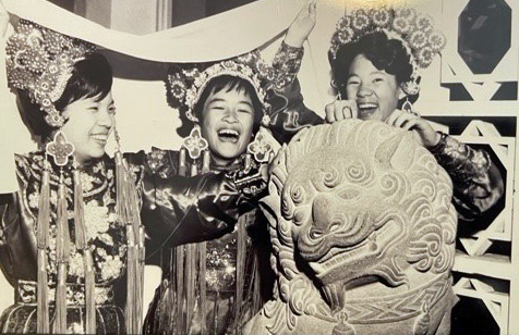 Black and white photo of girls breaking into laughter wearing Chinese traditional costumes and leaning on a lion sculpture.