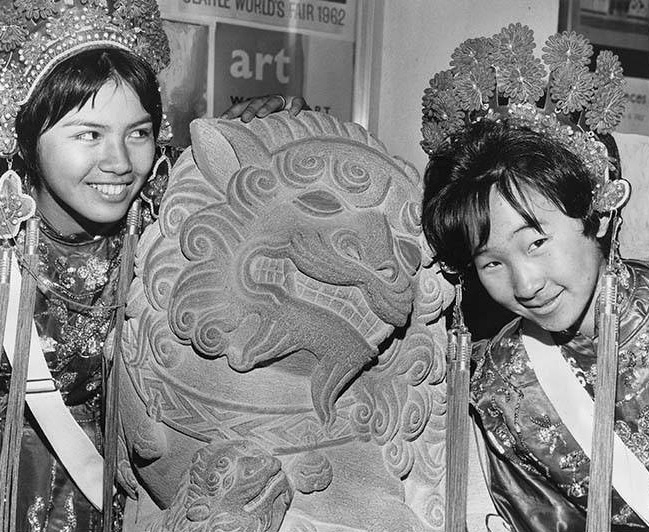 Black and white photo of two young women wearing Chinese Girls Drill Team headdresses, smiling and having fun with a lion sculpture between them.