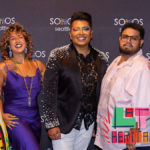 Five people, smiling, and dressed for a celebratory occasion standing together in front of a black backdrop with the logo for Somos Seattle printed on it.