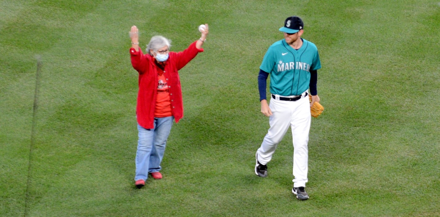 woman in red coat and jeans stands on baseball field with a player in blue Seattle Mariners uniform. her arms are raised upwards and she is holding a baseball in her left hand.