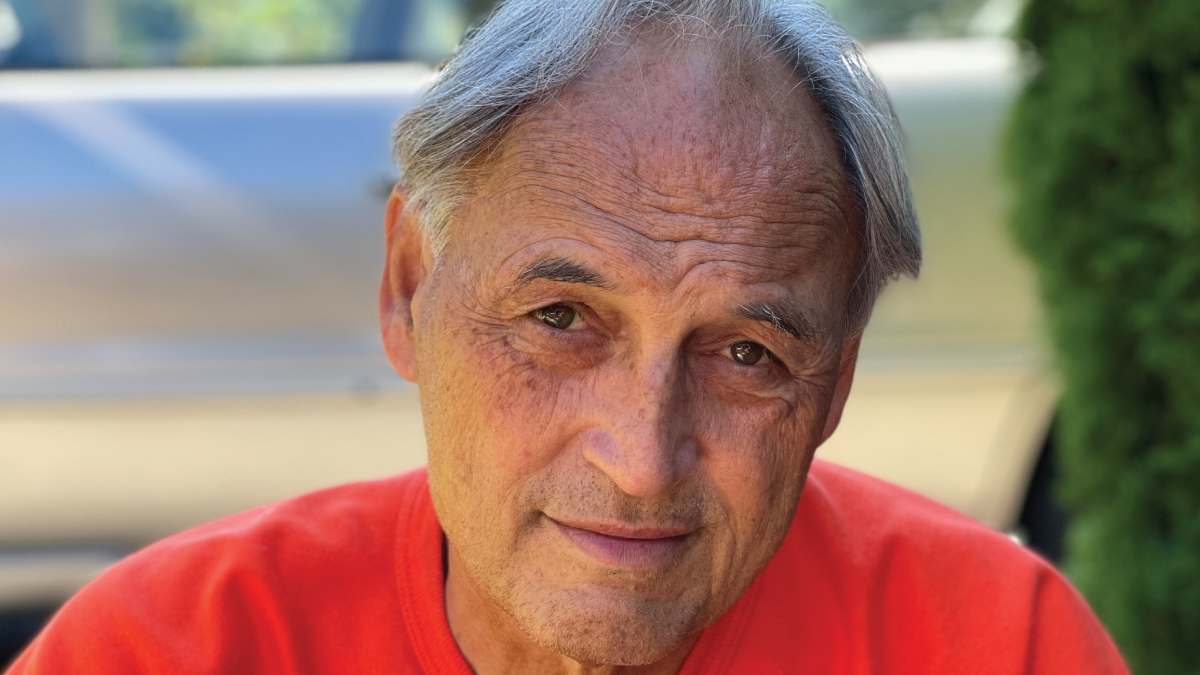 closeup of Native American man's face looking directly into camera. he has grat hair and is wearing a red tshirt. he is smiling warmly.