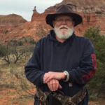 Native American man with white beard standing in desert location. he is wearing a sweatshirt and hat.
