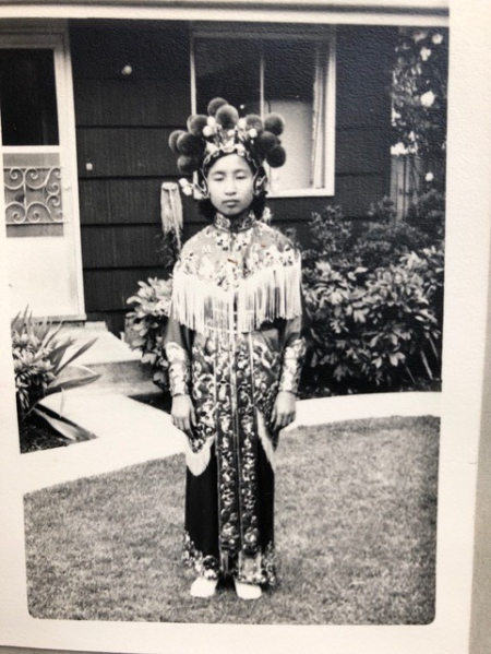 Young Betty Lau stands in front of a well kept house on a trimmed lawn wearing traditional Chinese Drill Team costume complete with shoulder fringes, pompom headress and silk brocade gown with a very serious look on her face.