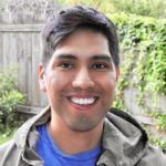 Native American man with short, dark hair, blue t-shirt and brown jacket. he is standing outside and smiling toward camera.