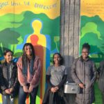 Six teenage girls standing in front of a colorful mural with text that reads "Our space will be a little different"