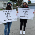 two masked young people standing in neighborhood road with stenciled signs that have the image of a salmon with the words "Safe River, Safe Salmon" and "Rio Seguro, Salmon Seguro"