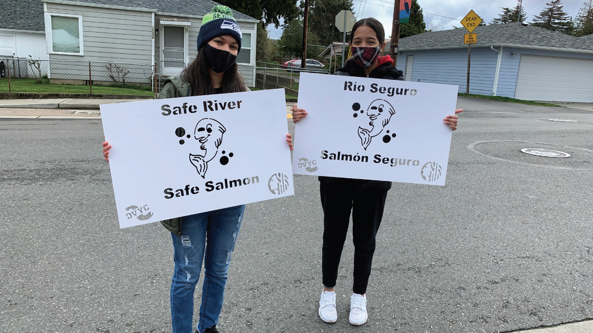 two masked young people standing in neighborhood road with stenciled signs that have the image of a salmon with the words 
