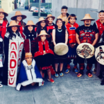 Group of Haida people in performance costumes and traditionally woven basket hats, holding painted drums.