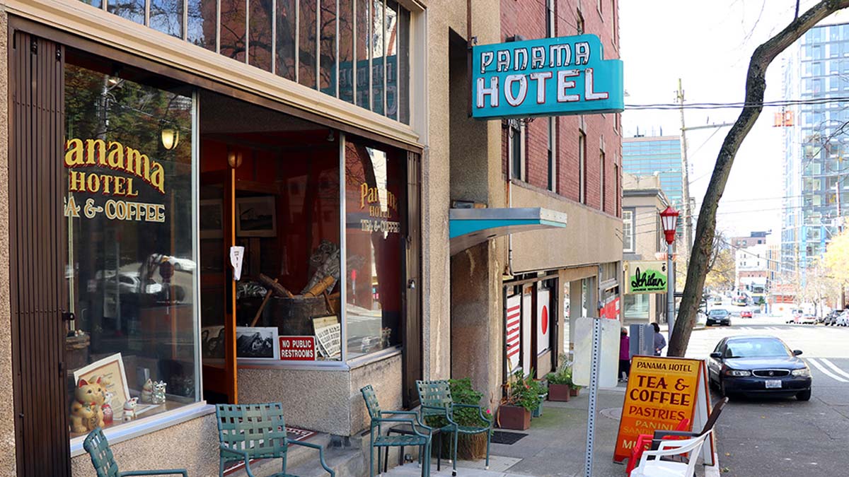 Cute little storefront with tall windows and metal chairs and tables on the sidewalk. Historical neon sign reads Panama Hotel against a turquoise blue background.
