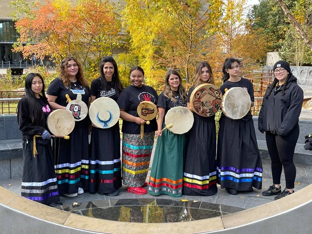 group photo of eight Native women wearing ribbon skirts and holding handmade drums. . They are standing outside of the National Museum of American Indian.