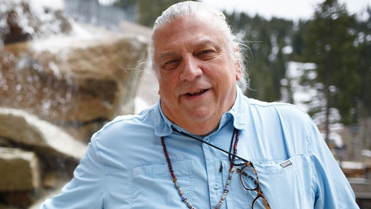 Indigenous man with long white hair stands outside in front of a fountain. he is smiling, wearing a blue button up shirt with glasses tucked into the pocket.