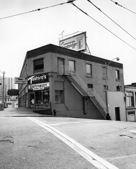 Black and white photo from the crosswalk of the corner entrance to Tashiro Kaplan Hardware with signs reading, Tashiro's Bait and Tackle, Outboard Shop, Johnson authorized dealer and a Cambridge cigarettes billboard perched on the top advertising, "Lowest tar ever."