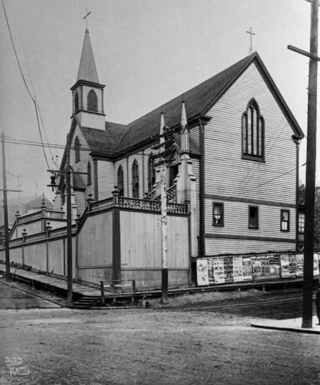 Black and white photo of a wooden church with powerlines coming from nearby poles and attaching to the structure, surrounding fence covered in posters or bills. The church is ringed by a wooden, elevated boardwalk above a muddy dirt street.