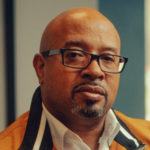 face of Dominique Davis, a bald Black man with goatee and glasses. his expression is serious.