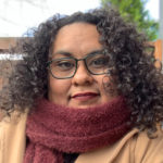 face of Eileen Jimenez. she has long, dark, curly hair. she is wearing glasses and has a red scarf wrapped around her neck.