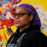 Black woman wearing tinted glasses looks directly at the camera. she has a bandana tied around her head and is wearing pink lipstick. she is standing in front of a brightly painted mural.