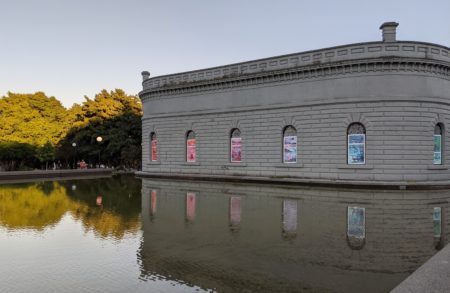 Photo of a gray building with castle-style architectural with five windows featuring  brightly colored, illustrated art pieces snugly fit into the  frames. A reflecting pond surrounds the structure with leafy trees in the distance reflected in the water. 