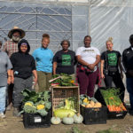 A group of farmers and volunteers at Clean Greens Farmstand in front of a large greenhouse with freshly harvested vegetables displayed in boxes.