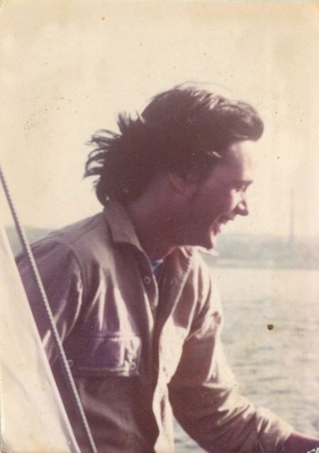 An aged photograph close up of young man with dark hair blowing in the wind - Rob Wallace - on a sailboat on the water, smiling, face in profile, and wearing a flannel button up.
