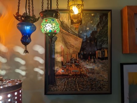 Photo of an oil-painting depicting a terrace on a cobblestone street with evening light, stars. The painting is lit by three, retro hanging pendant lights of different colored glass. 