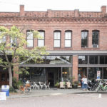Brick storefronts on old Ballard Avenue with cafe tables, people drinking coffees, bikes parked out front and beautiful tree providing dappled shade