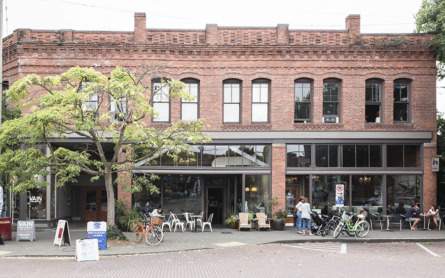 Brick storefronts on old Ballard Avenue with cafe tables, people drinking coffees, bikes parked out front and beautiful tree providing dappled shade