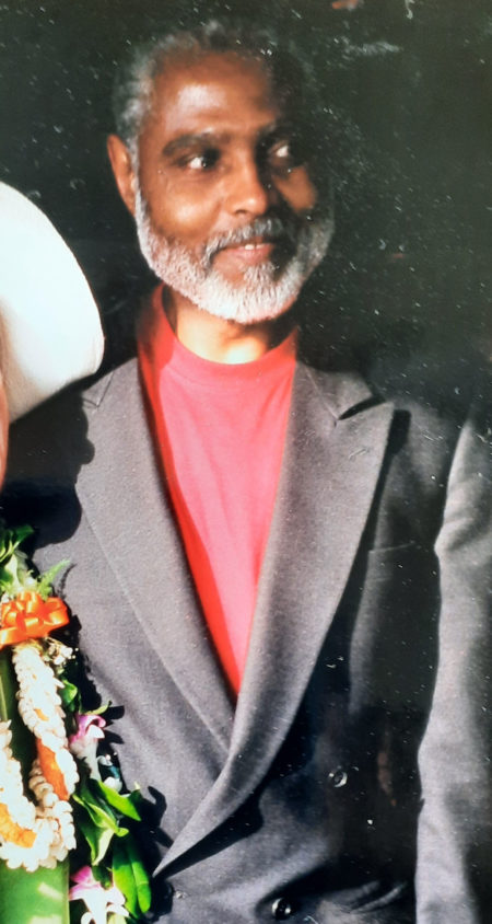 Black man with gray hair and beard, smiling. He is wearing a dark suit jacket over a red t-shirt.