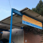 A blue and yellow sign that reads "Sacajawea" hangs over a covered walkway into a brick building