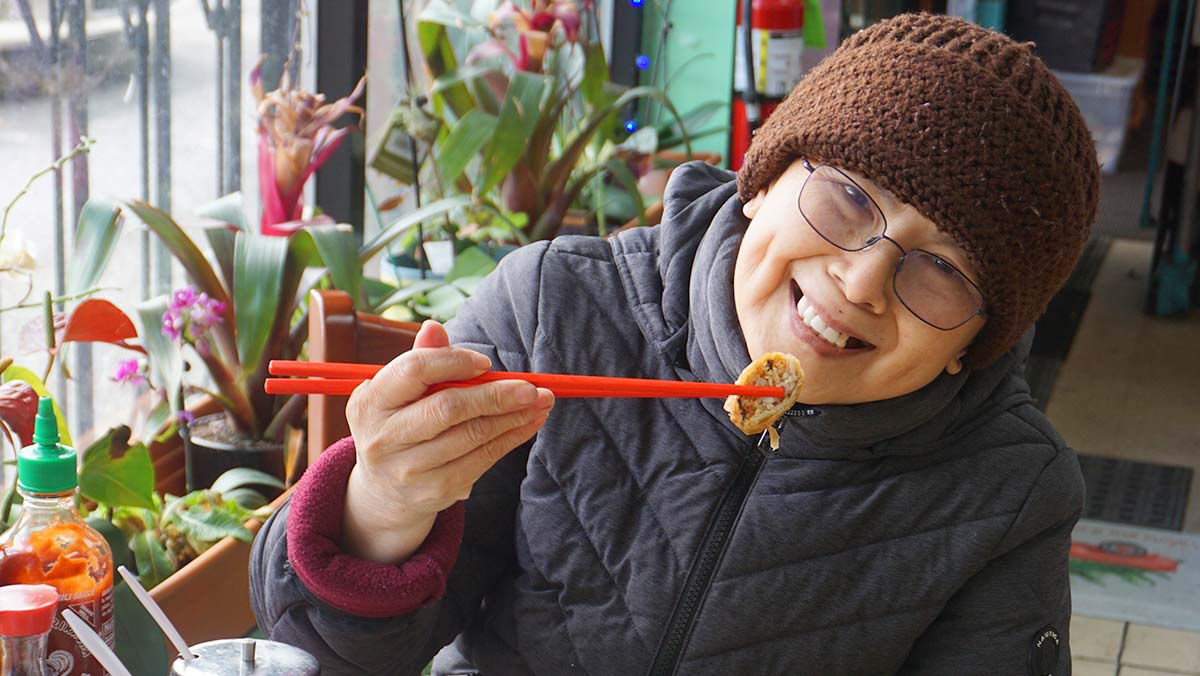 An Asian woman wearing a black hat and brown coat smiles while holding up a piece of tofu with chopsticks