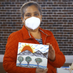 A person wearing a protective facemask holding up a small canvas painting with trees, mountains, and the sun