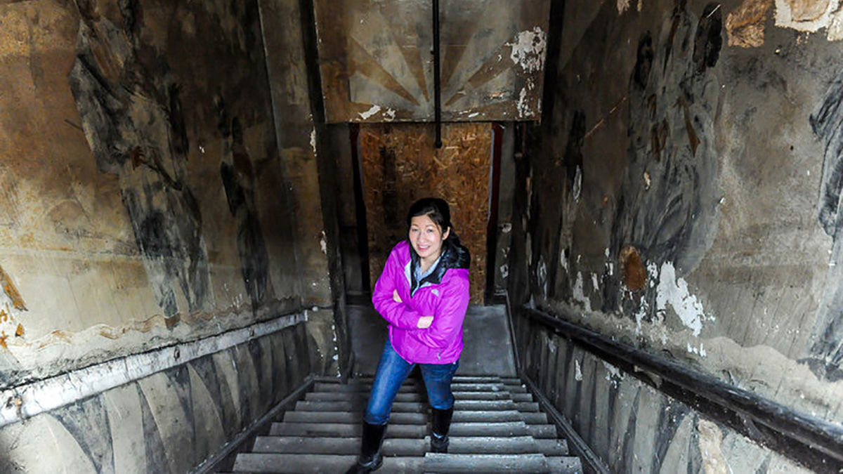 An Asian American woman standing down in a stairwell with arms crossed looking up at the camera
