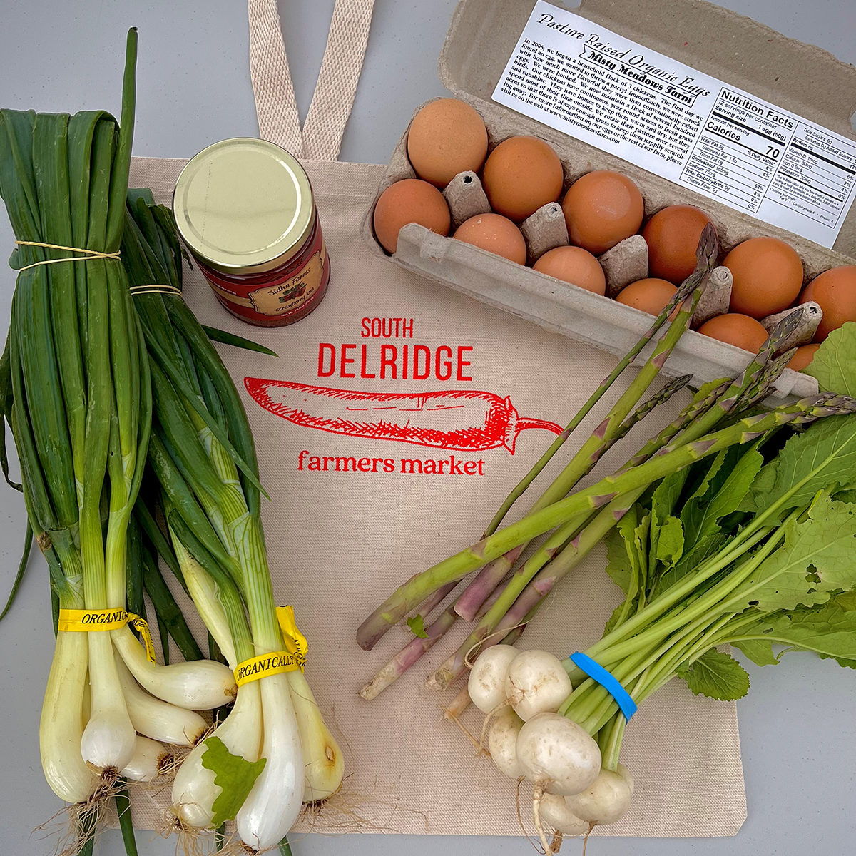 carton of brown eggs, produce, and a jar of jam sitting on top of a cloth bag that reads "South Delridge Farmers Market"