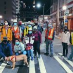 A group of people wearing high visibility reflective vests and safety masks stand in a city crosswalk at night