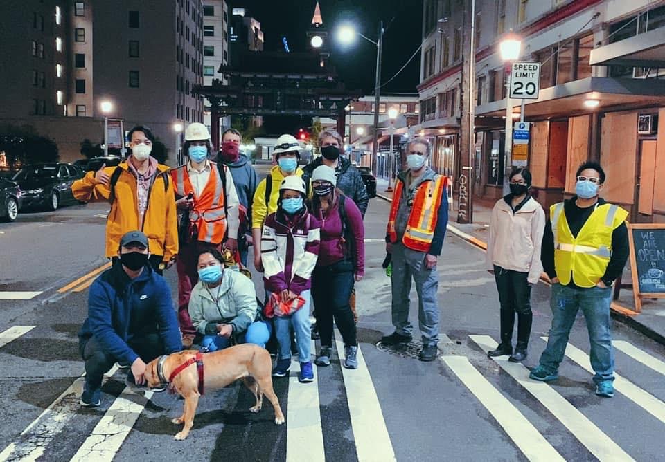 A group of people wearing high visibility reflective vests and safety masks stand in a city crosswalk at night