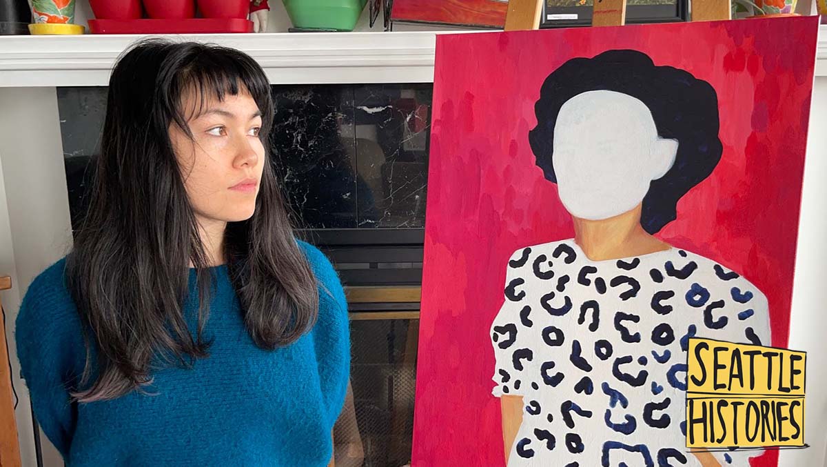 A Taiwanese American woman standing beside and looking at a painting of a person with the face whited out.